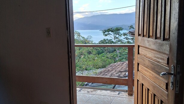 An open wooden door leading to a balcony with a tiled floor and a view of a body of water and mountains. - A house to enjoy and take advantage of beaches, waterfalls, and many adventures in Ilhabela. (Ilhabela)