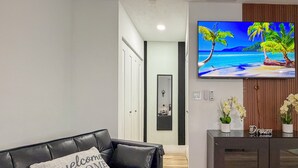 A modern living room with a black leather sofa, a TV displaying a beach scene, a wooden cabinet, and a 'Welcome Home' pillow. - Modern Luxury Studio • Kitchen, Wi-Fi & Private Parking (Pembroke Pines)