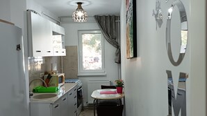 Een compacte keuken met witte kastjes, een ingebouwde magnetron, en een gootsteen. Er staat een eettafel met stoelen en een potplant. Een raam met gordijnen is zichtbaar, en er is een plafondlamp.