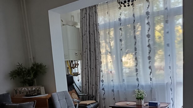 Een woonkamer met een grijze bank, een zwart-wit kleed, een houten salontafel en een stoel. Er is een groot raam met dunne gordijnen en uitzicht op bomen.