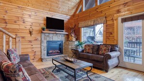 L’intérieur chaleureux d’un chalet avec une cheminée en pierre, un canapé en cuir et un téléviseur.