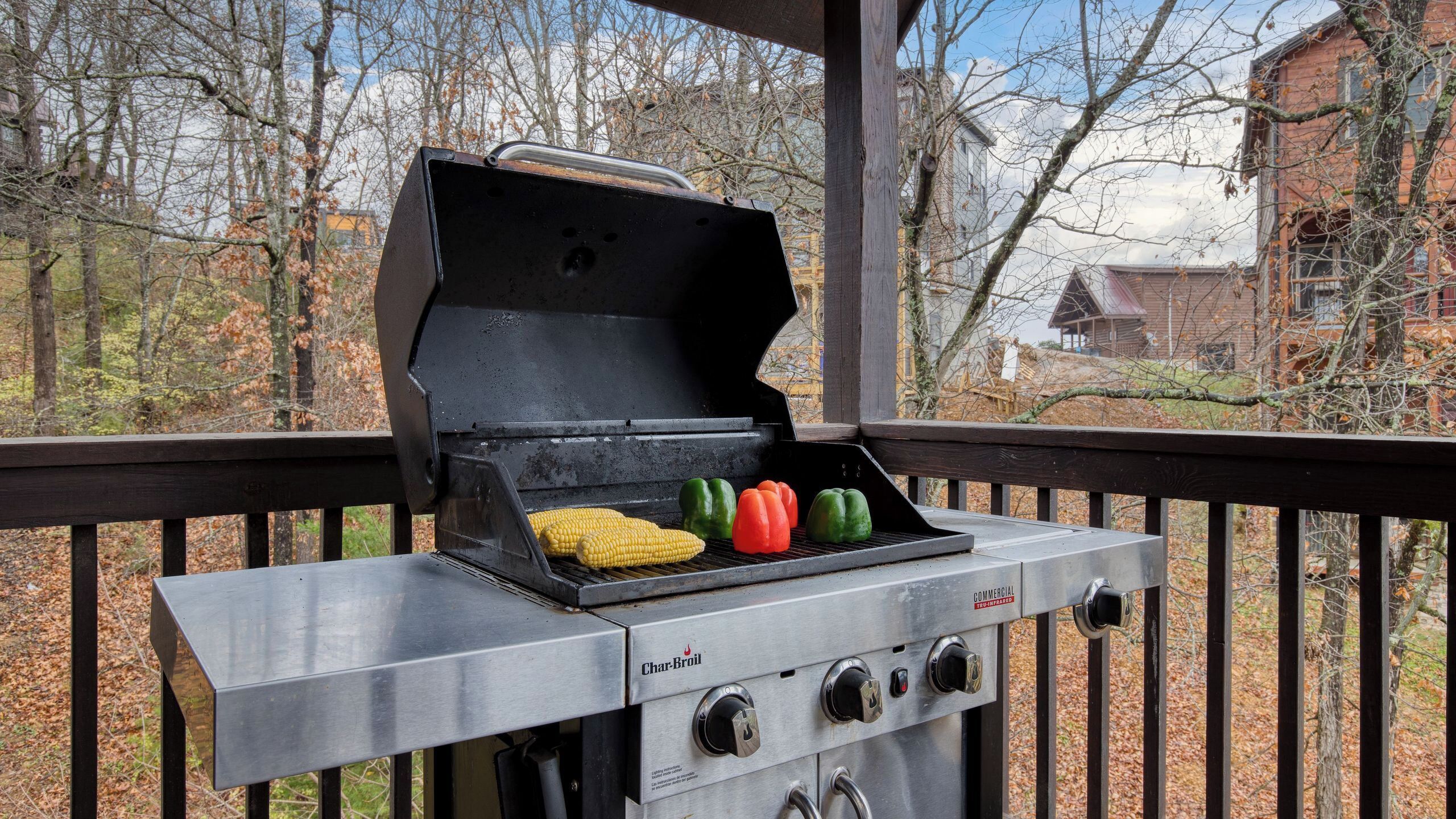 Un barbecue Char-Broil avec des légumes sur la grille, une table en métal et une terrasse en bois offrant une vue sur les arbres et les bâtiments.