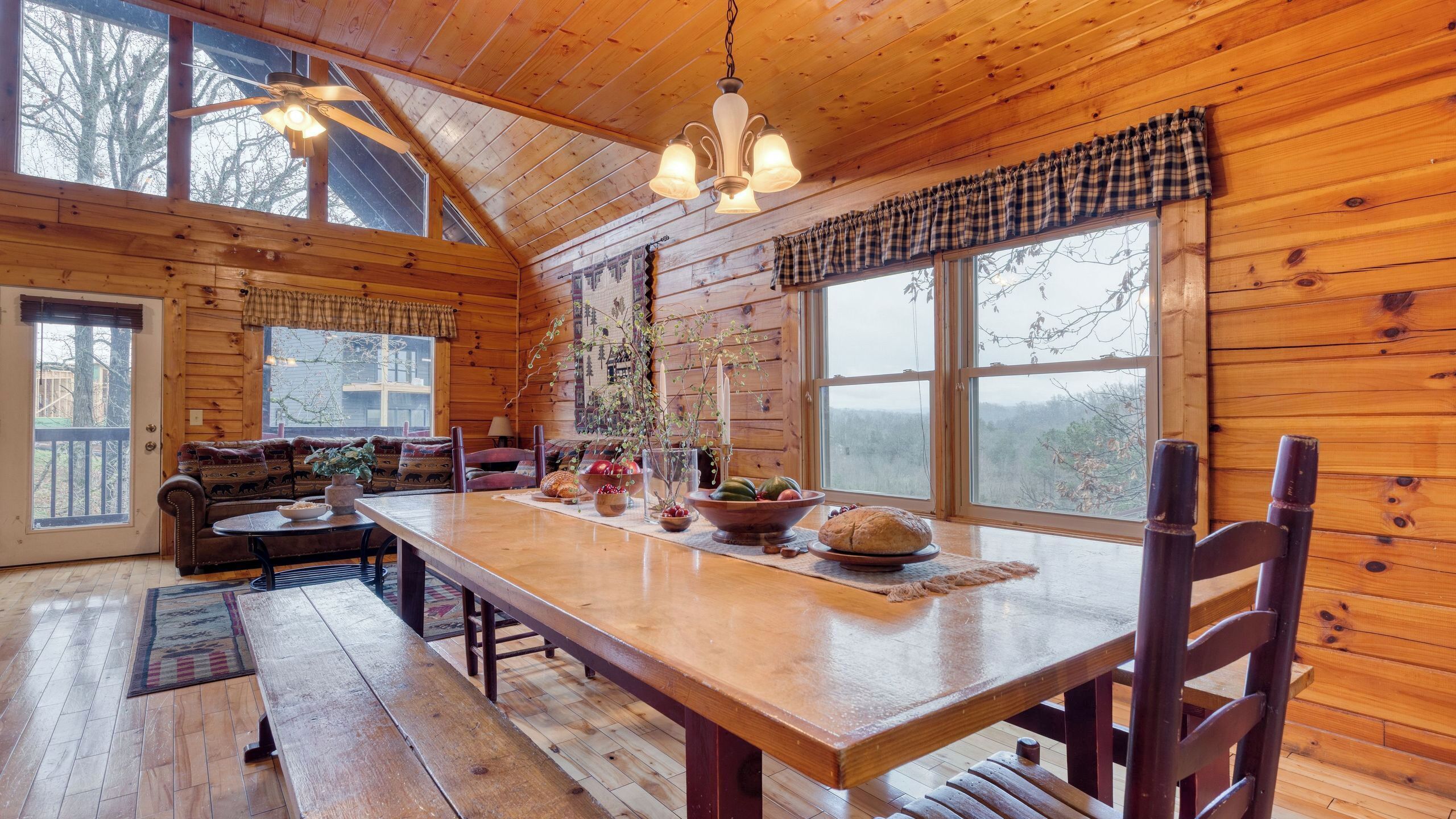 A cozy cabin interior with a wooden dining table, benches, a sofa, and large windows offering views of the outdoors.