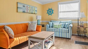 A room with a yellow wall, a painting of beach huts, a striped sofa, an orange sofa, a small table, a ship's wheel decoration, and a window with blinds. - BeachTimes and TanLines|GolfCart|Linens|OceanLakes (Horry County)