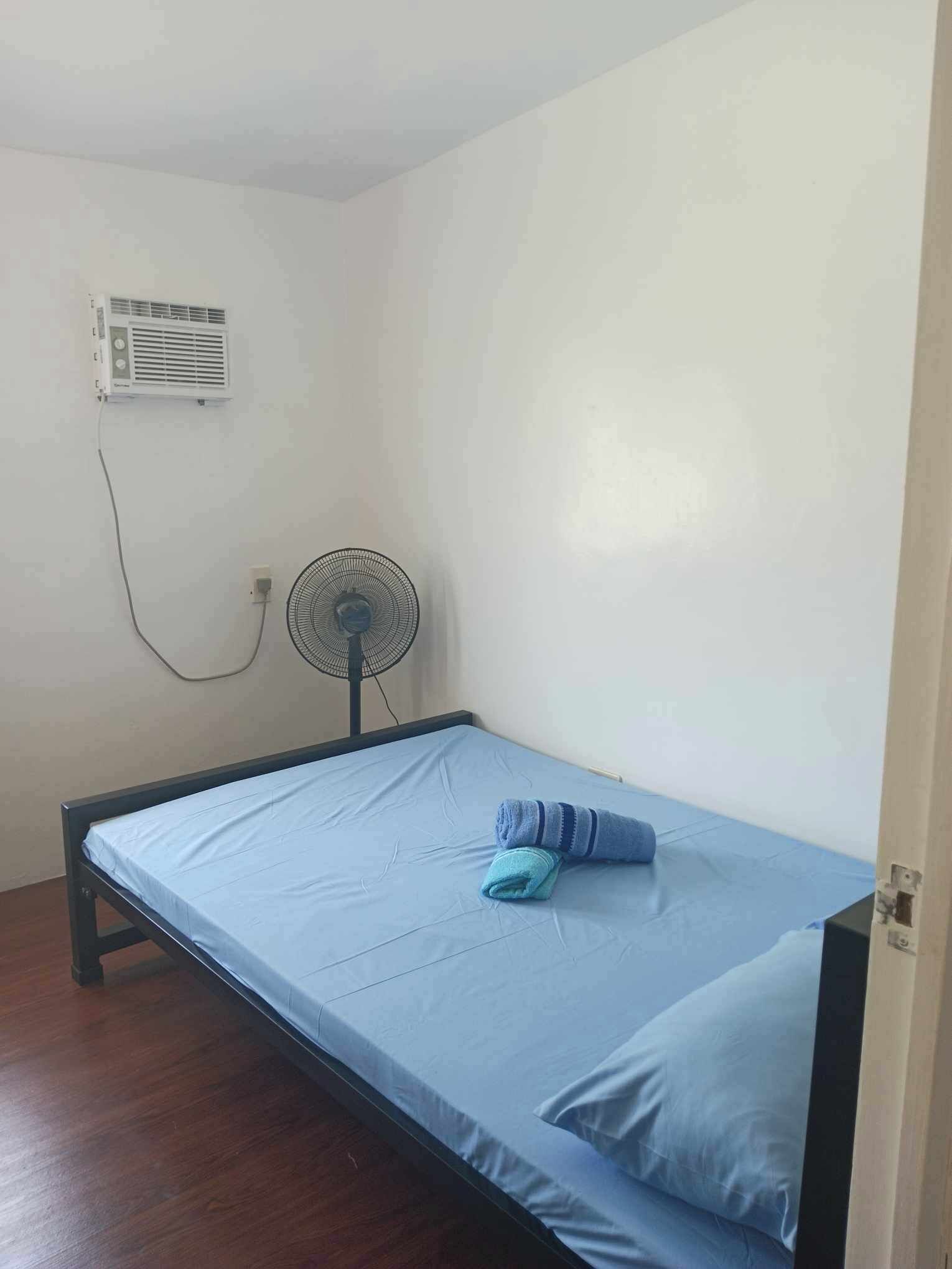 A single bed with a blue mattress and a matching pillow, a standing fan, and a wall-mounted air conditioner in a simple room with white walls and wooden flooring.