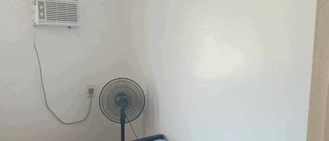 A single bed with a blue mattress and a matching pillow, a standing fan, and a wall-mounted air conditioner in a simple room with white walls and wooden flooring.