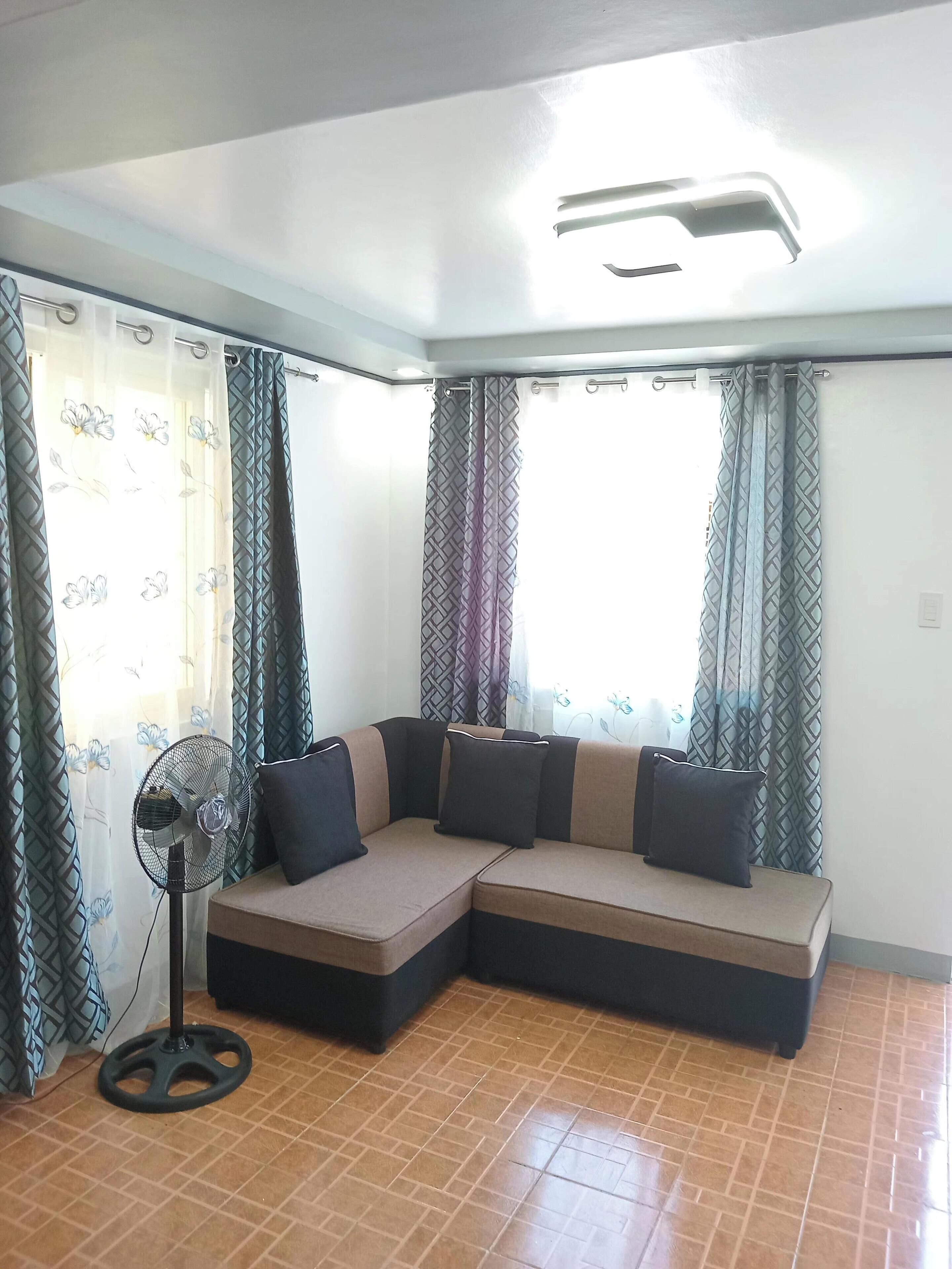 A corner room with a sectional sofa, a standing fan, and patterned curtains.