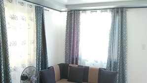 A corner room with a sectional sofa, a standing fan, and patterned curtains.