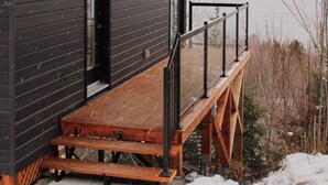A modern cabin with a wooden deck and a glass door, overlooking a lake and mountains. - Refuge du Fjord (Saguenay)