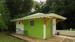 Un piccolo edificio a un piano con facciata verde e porta bianca, dotato di un patio con pavimento in pietra e un tavolino con una sedia.