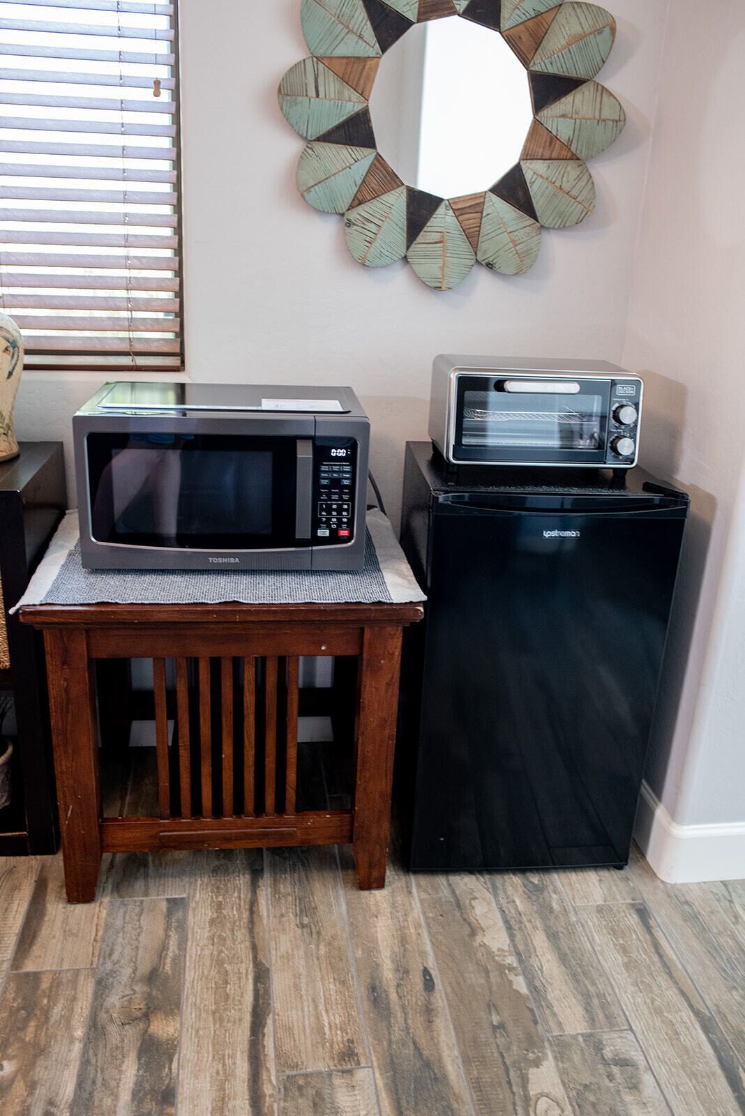 Een keuken met een magnetron en een broodrooster op een kleine houten tafel, een decoratieve spiegel aan de muur, en een raam met jaloezieën.