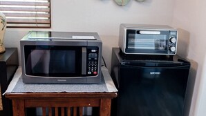 Een keuken met een magnetron en een broodrooster op een kleine houten tafel, een decoratieve spiegel aan de muur, en een raam met jaloezieën.