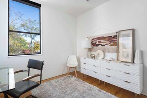 Ein modernes Zimmer mit einem Glastisch, einem schwarzen Stuhl, einer weißen Kommode und einem großen Fenster mit Blick auf Bäume.