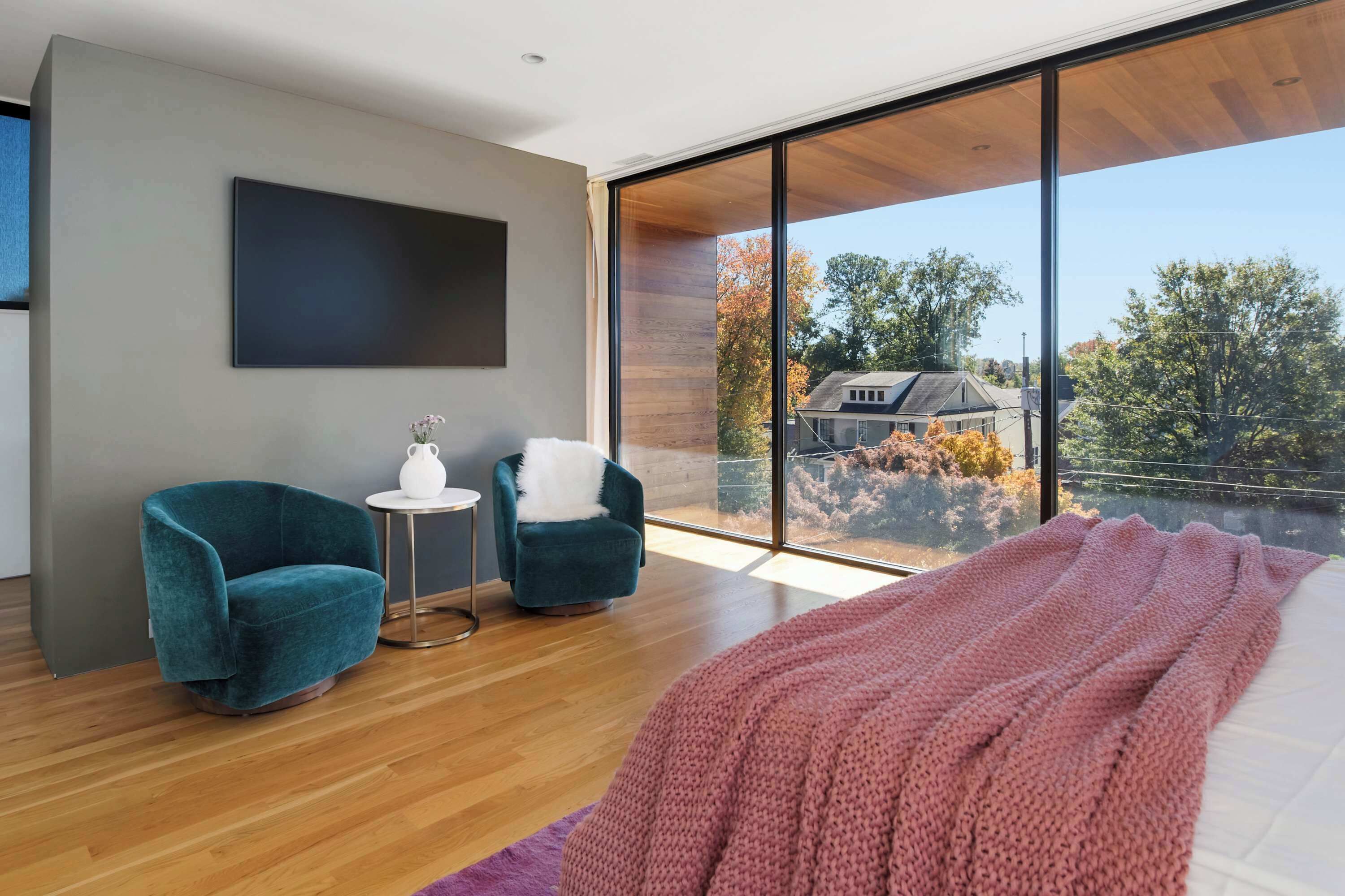 A modern bedroom with a large window, two chairs, a small table, and a bed with a pink blanket.