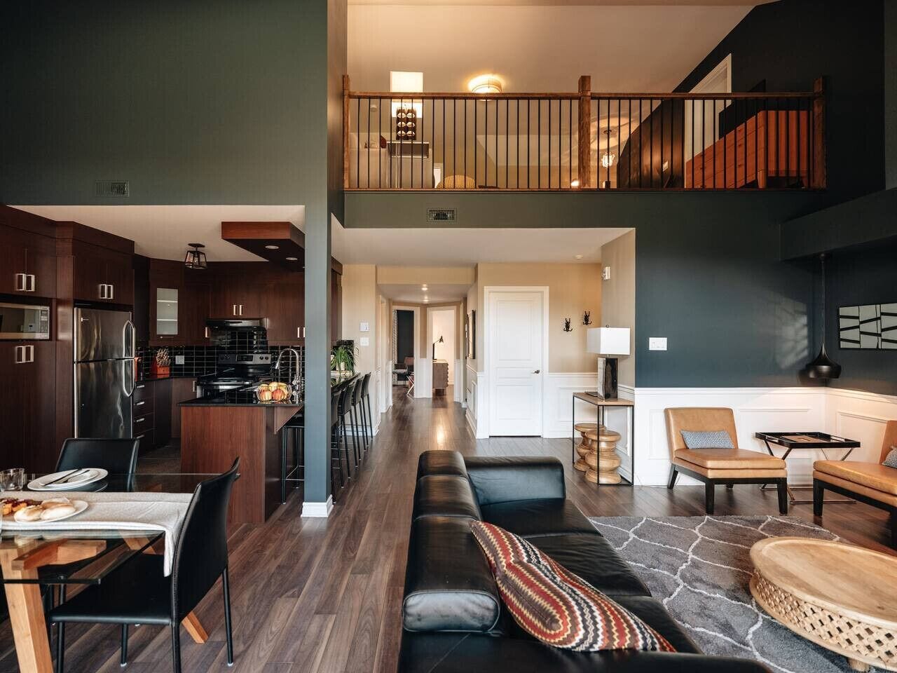Une cuisine moderne avec des meubles en bois foncé et des appareils électroménagers en acier inoxydable, un coin repas avec une table dressée pour deux personnes, et un espace salon confortable avec un canapé et des fauteuils.