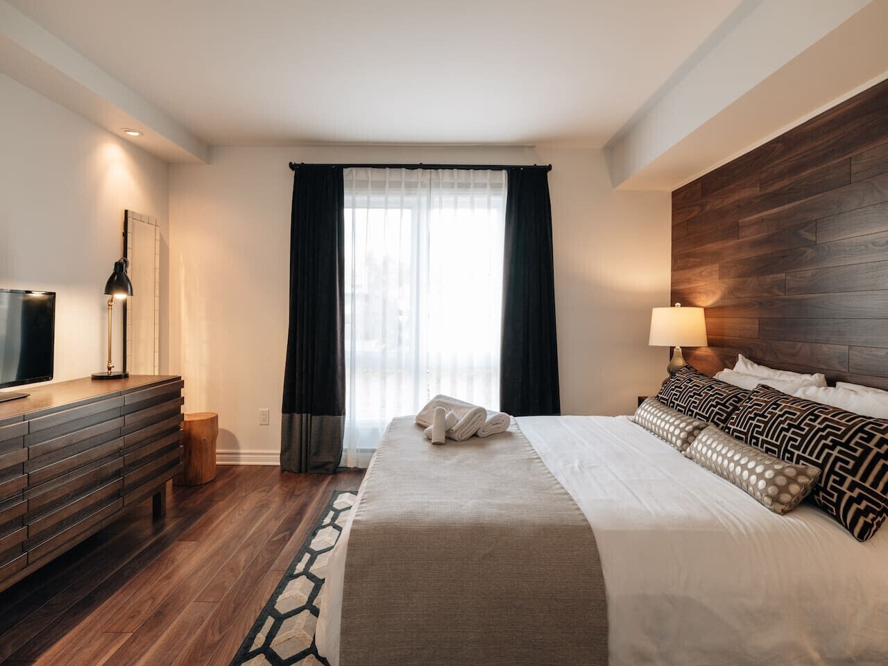 A hotel room with a bed, a dresser, a TV, and a window with curtains.