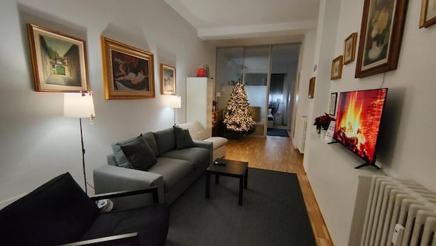 A living room with a grey sofa, a black chair, a small table, a Christmas tree, and a TV on the wall. - Short Rent Milan Porta Romana  (Milan)