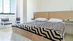 Un dormitorio con cama, mesita de noche, una silla y vistas a un paisaje urbano desde un balcón.