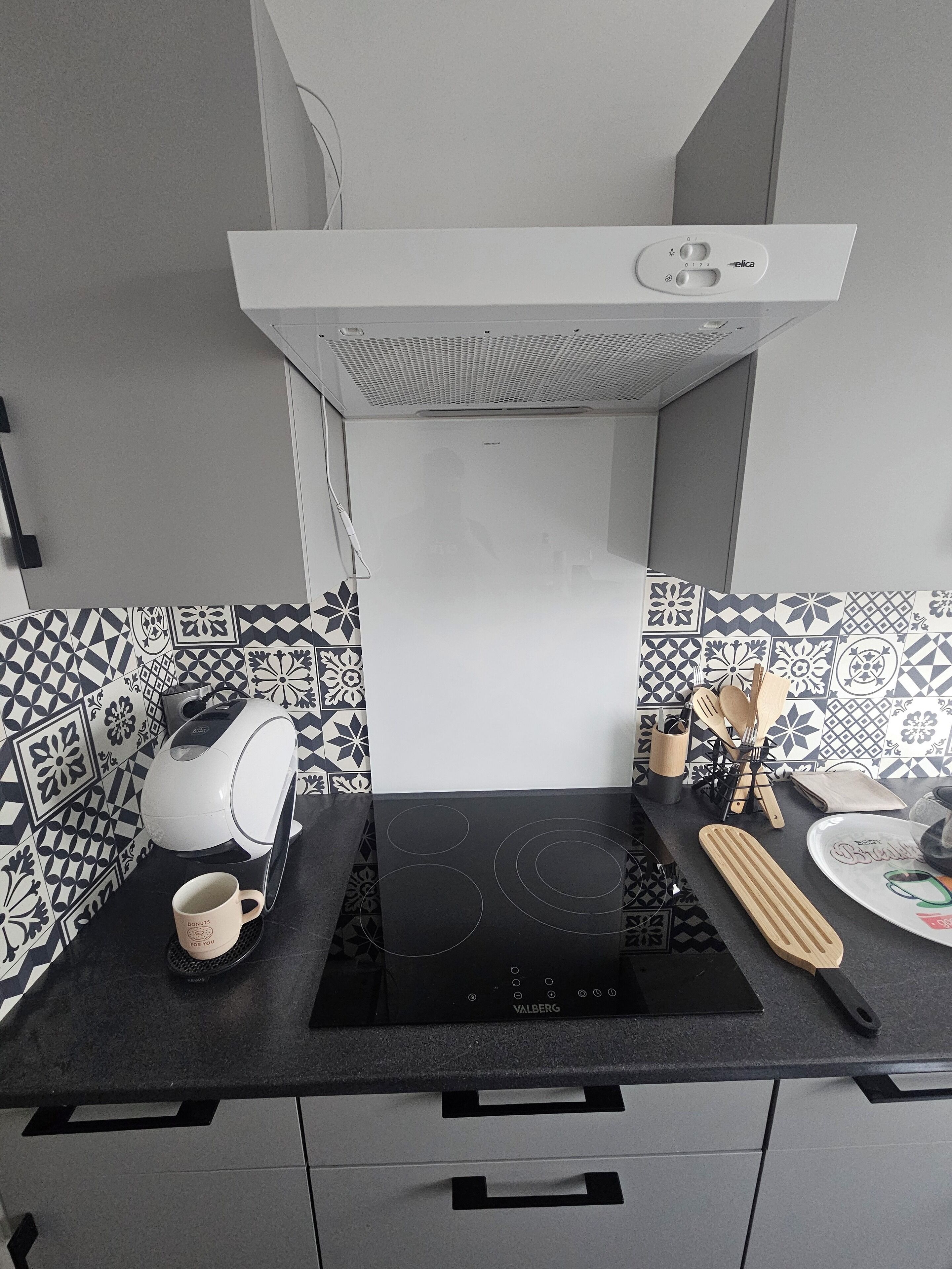 Une cuisine dotée d’un plan de travail noir, d’une bouilloire blanche, d’une tasse à café, d’une plaque de cuisson à induction noire et d’un pare-feu orné de motifs géométriques.