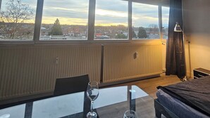 Une chambre d’hôtel moderne avec une grande fenêtre, une table en verre avec deux verres à vin, un lit et une lampe de sol.