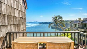A balcony with a wooden table and wicker chairs, overlooking a coastal view with palm trees and a building with solar panels. - Oceanfront Eagle’s Nest - Bali Hai Sunset Views (Princeville)