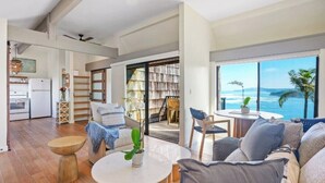 A spacious living area with a large window offering a view of the ocean, a cozy seating area with a coffee table, and a kitchen in the background. - Oceanfront Eagle’s Nest - Bali Hai Sunset Views (Princeville)