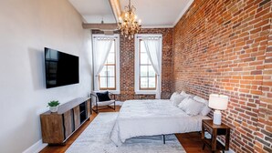Ein Schlafzimmer mit Backsteinwand, Flachbildfernseher, Bett, Nachttisch, Lampe und Stuhl.