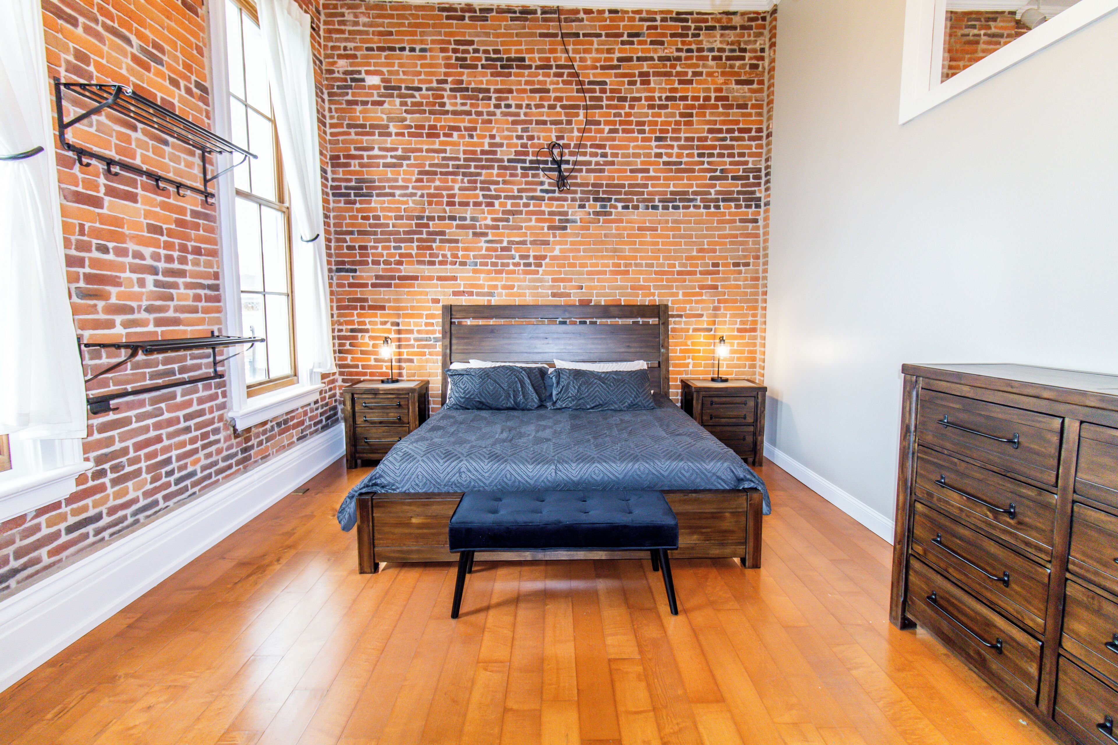 A bedroom with a brick wall, wooden floor, a bed with a headboard, two bedside tables, and a dresser.