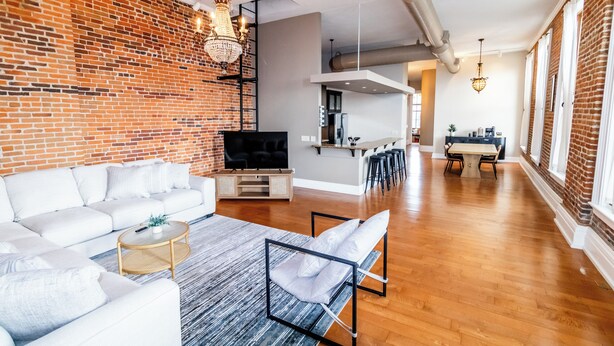 Ein modernes Wohnzimmer mit einem Ecksofa, einem Couchtisch und einem Fernseher. Im Hintergrund befindet sich ein Küchenbereich mit Barhocker und einem Esstisch.