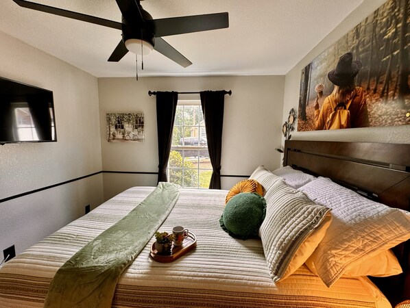 A bedroom with a bed, a ceiling fan, a window with curtains, a painting on the wall, and a small table with a plant and a cup. - Hot Tub • Firepit 3BR Bike-in/out Nature Home (Bella Vista)