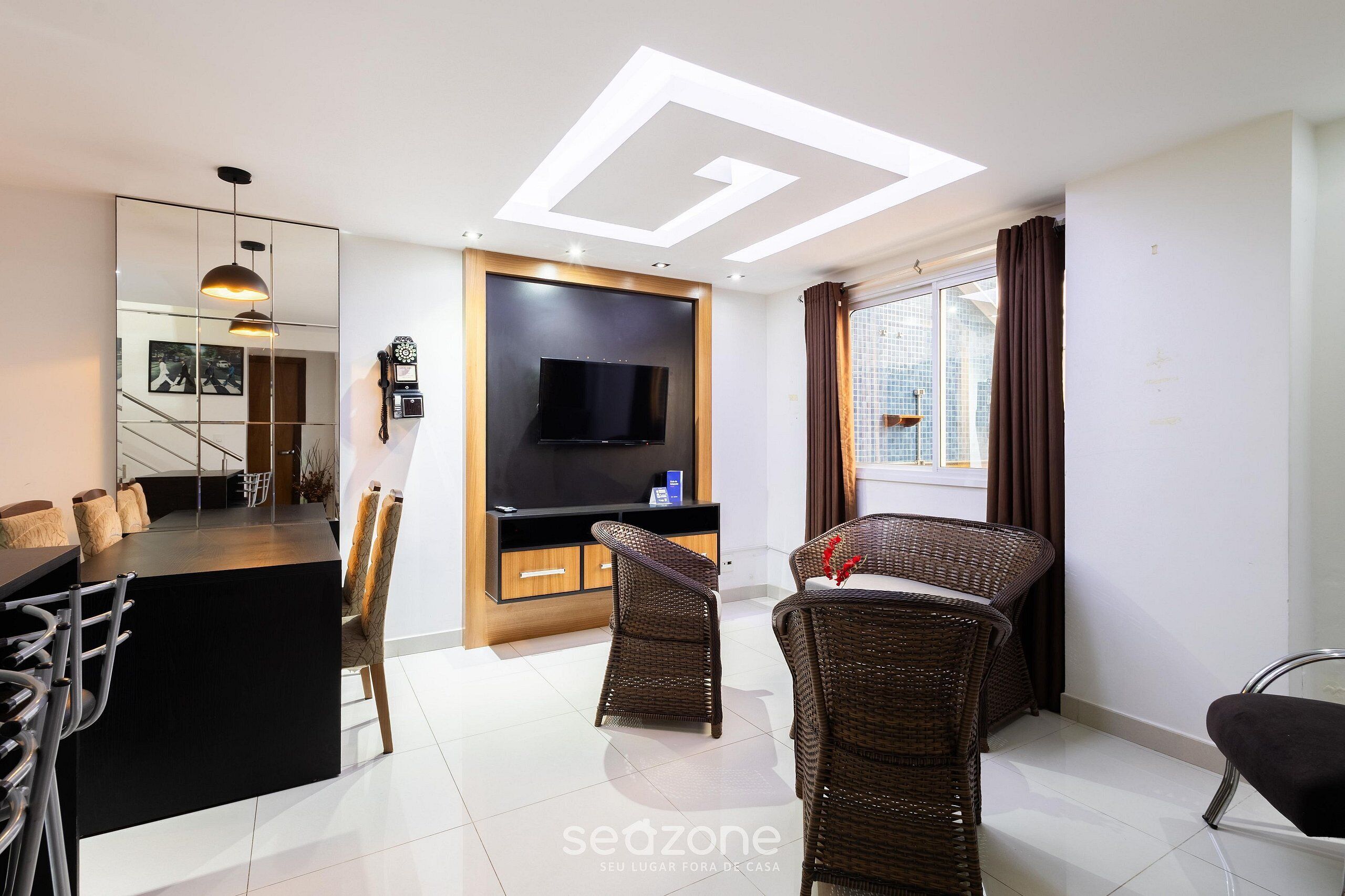 A modern living room with a flat-screen TV, wicker chairs, a bar area, and a large window with curtains.