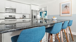A modern kitchen with white cabinets, a stainless steel microwave, and a granite countertop. There are blue bar stools and a framed picture of a palm tree on the wall. - Pool, Hot Tub, Pickleball, Game Room & Spacious for Groups - Near Beach! (Gulfport)