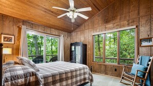 Une chambre à coucher située dans une cabane en bois, avec un lit, un fauteuil à bascule et de grandes fenêtres donnant sur la forêt.