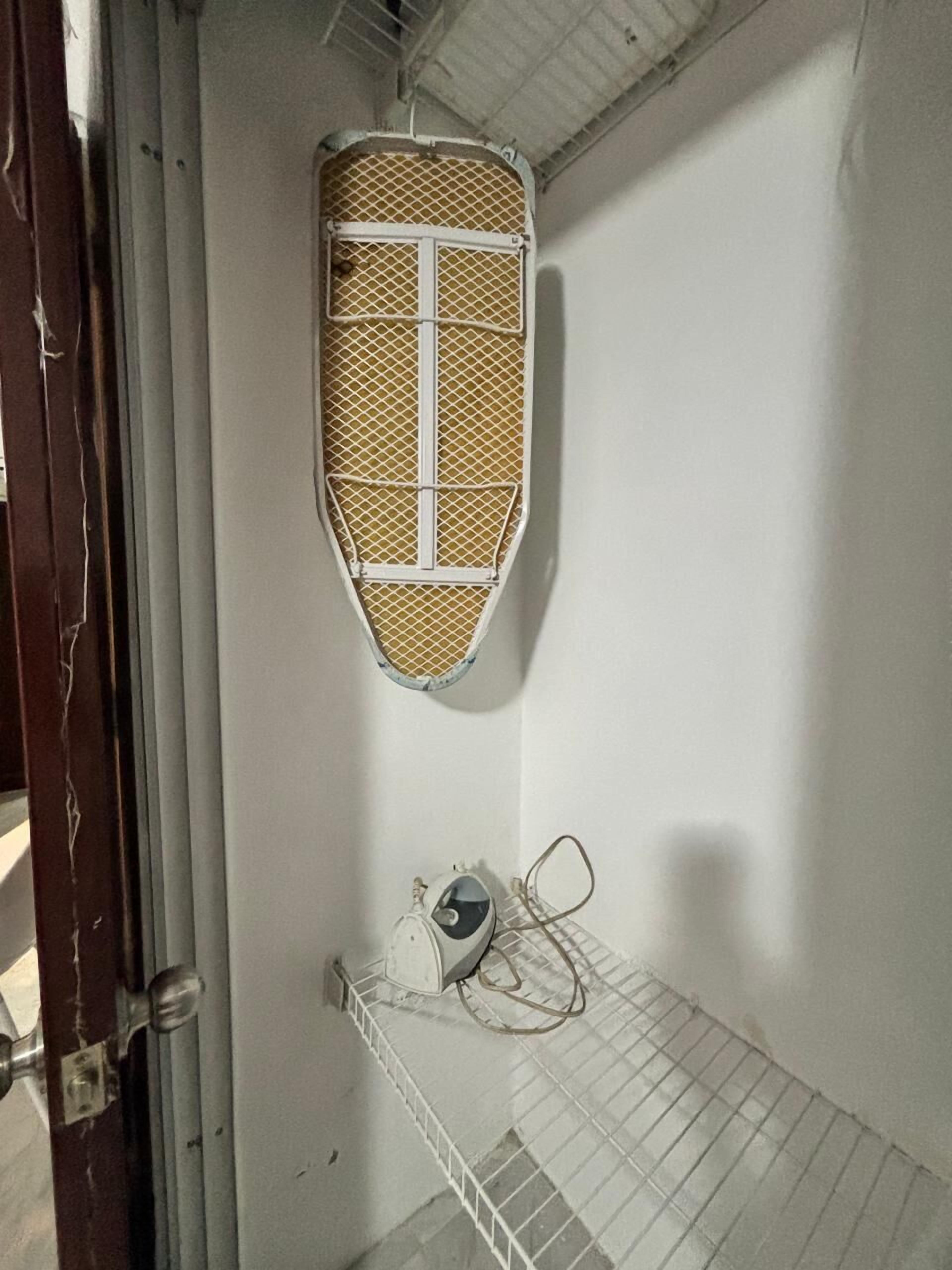 A room with a metal shelf, a wall-mounted ironing board, and a door with a metal handle.