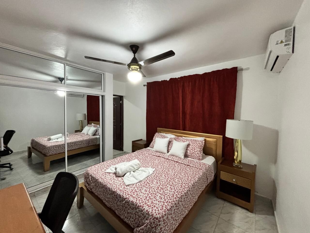 Ein Hotelzimmer mit einem Bett, einem Schreibtisch, einem Stuhl, einem Deckenventilator, einem Spiegel und einer Klimaanlage.