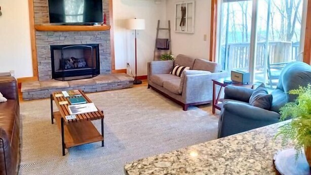 A living room with a stone fireplace, a flat-screen TV mounted above it, a grey sofa, a coffee table, and a large window with a view of trees. - Modern comfort with mountain charm! (Mars Hill)