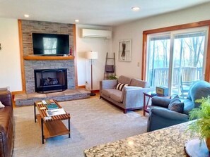 A living room with a stone fireplace, a flat-screen TV mounted above it, a grey sofa, a coffee table, and a large window with a view of trees. - Modern comfort with mountain charm! (Mars Hill)