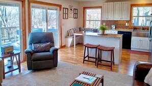 A spacious living area with a large window, a comfortable sofa, a small coffee table, and a kitchen area with white cabinets and a wooden island. - Modern comfort with mountain charm! (Mars Hill)