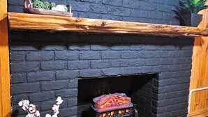 En öppen spis med en svart tegelstensdesign, en trähylla ovanför och en vas med vita blommor.
