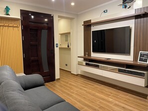 A modern living room with a grey sofa, wooden flooring, a large TV on a wooden stand, and a door with a mirror. - GAFI 2 Lodging - Main Avenue (Independencia)