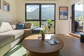 Ein Wohnzimmer mit einer Couch, einem Couchtisch mit einer Zeitschrift und Blick auf Berge durch große Fenster.