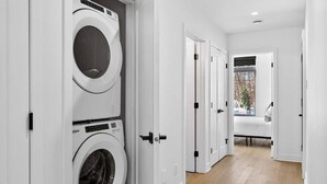 A laundry room with two stacked washing machines, a bench, and a window with a view of trees. - The Axia at Verbier: Hot Tub, Sauna, Seasonal Pool (Mont-Tremblant)