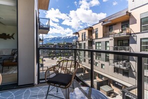 Ein Balkon mit einem Korbstuhl, Blick auf ein modernes Gebäude mit mehreren Balkonen und ein Gebirge im Hintergrund.