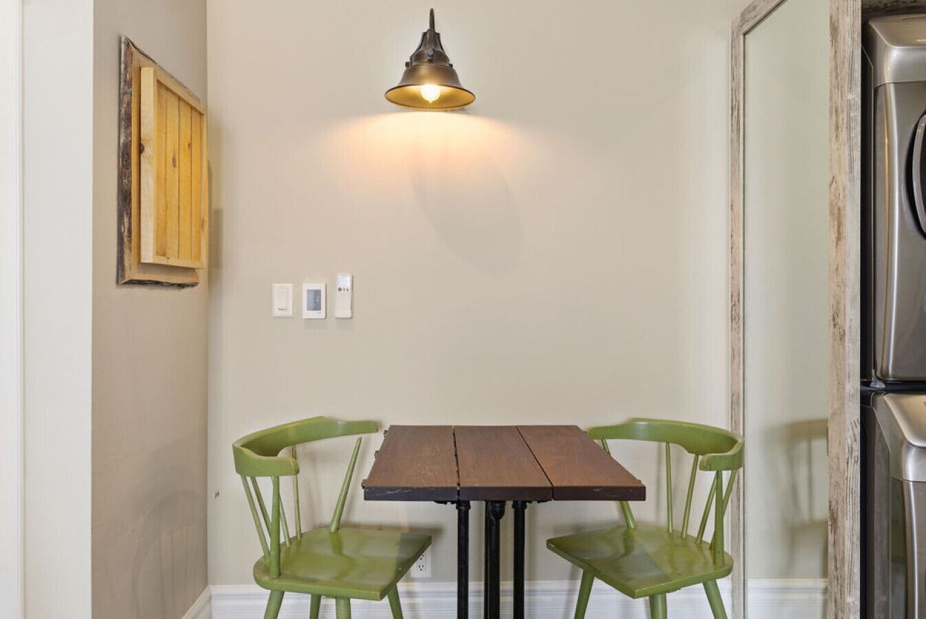 Een eethoek met een houten tafel en twee groene stoelen, een hanglamp en een schilderij aan de muur.