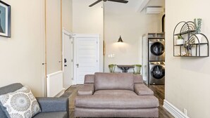Un salon comprenant un canapé, une table basse et un coin repas avec des chaises. On y trouve également un ventilateur de plafond, une étagère murale ornée de plantes et une porte donnant sur une autre pièce.