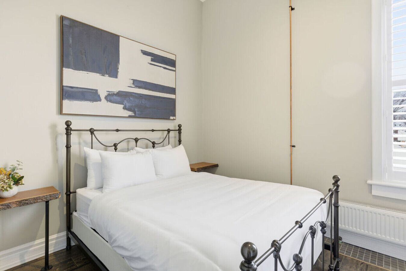 A neatly made bed in a room with a simple metal headboard, a bedside table with a vase of flowers, and a large abstract painting on the wall.