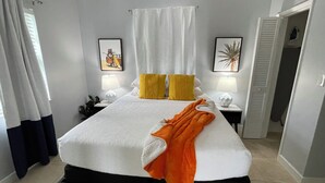 Una cama bien hecha con edredón blanco, dos almohadas amarillas y una manta naranja. Hay dos mesitas de noche con lámparas, un cuadro enmarcado en la pared y una puerta que da a otra habitación.