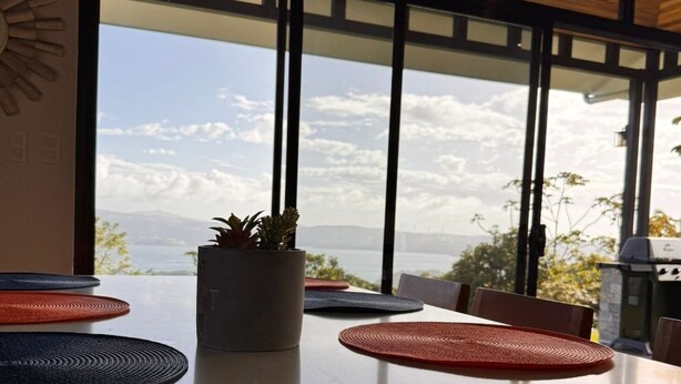 A modern dining area with large windows, a potted plant, and a view of the sea. - Lake Sunset House in Arenal-quiet retreat spectacular views (Tilaran)