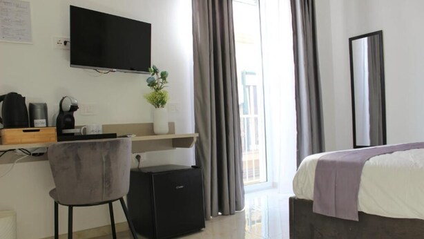 A hotel room with a bed, a desk with a chair, a small black refrigerator, a television mounted on the wall, and a window with curtains. - Cozy and equipped with amenities, for couples and families in a quiet setting. (Napoli)
