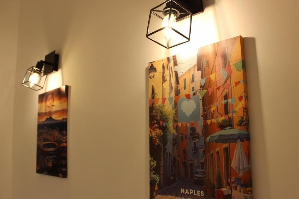 Deux œuvres d’art encadrées sont accrochées au mur : l’une représente un coucher de soleil et l’autre une scène de rue colorée, sur laquelle on peut lire le mot « NAPLES ».